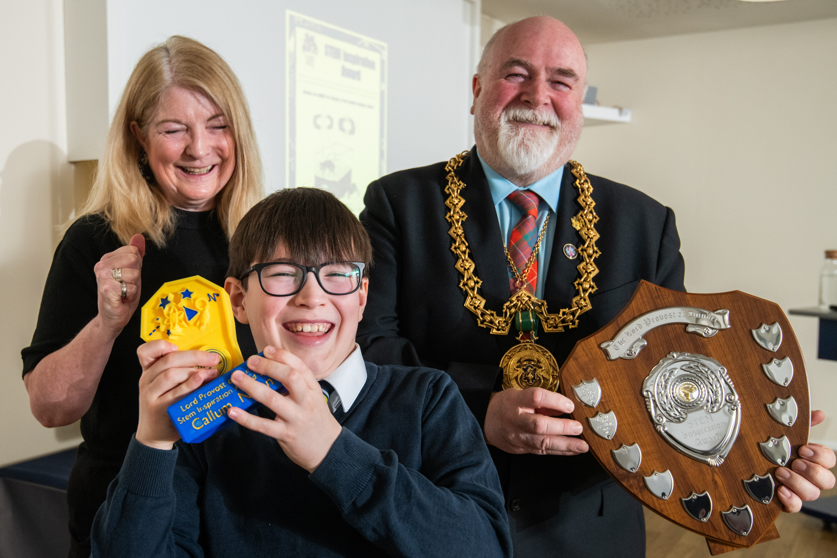 Callum, a school boy holds his award, he is joined by Jill the CEO of Dundee Science Centre, she’s smiling and clapping. Next to them, the Lord Provost holds a large shield shaped trophy.